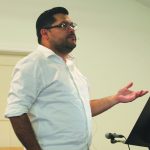 Man in white shirt speaking at a podium with a microphone during a presentation or lecture.