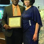 A man and woman smiling, holding a framed certificate at an indoor event.