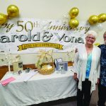 Elderly couple celebrating 50th anniversary with banner, balloons, and a decorated table with photos and cards.