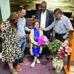 Group prays over girl with flowers in a church, standing near a floral arrangement and microphone stands.