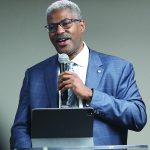 Man in a blue suit giving a speech with a microphone and tablet on a podium.