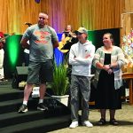 Church ceremony with a group of people on stage, one with a guitar and another holding a baby, in a wooden room.