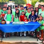Group of people celebrating outdoors with a cake on a blue table, wearing matching green shirts and smiling.