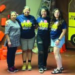 Four women posing together at a VBS event, wearing themed shirts, smiling in a decorated room.