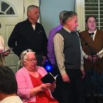 A group of people singing and holding notebooks in a room with microphones and a music stand.