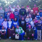 A large group of people gather outdoors for a cheerful family event in a rural setting, with a tractor in the background.