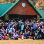 Large group of people posing in front of a cabin during fall, with colorful trees in the background.