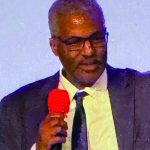 Man in a suit holding a red microphone speaks at an event.