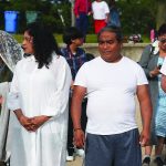 A group of people dressed in white standing outdoors at a gathering, with others in the background.