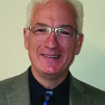 Man in glasses wearing a gray suit and blue tie, smiling against a light background.