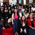 Group of people gathered indoors, smiling and standing together, some seated, in a community event or gathering.