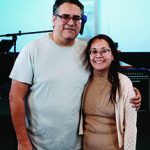 A happy couple standing together, smiling in a relaxed indoor setting, with musical equipment in the background.
