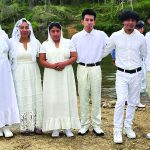 Group of people in white outfits standing together by a riverbank, surrounded by nature and greenery.