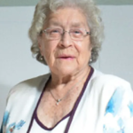 Elderly woman with glasses and white floral blouse, smiling softly against a neutral background.