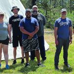 Group of nine adults smiling outdoors in front of a tent on a sunny day, wearing casual summer clothing.