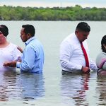 People participating in a water baptism ceremony at a lake, exchanging vows and holding hands in a serene setting.